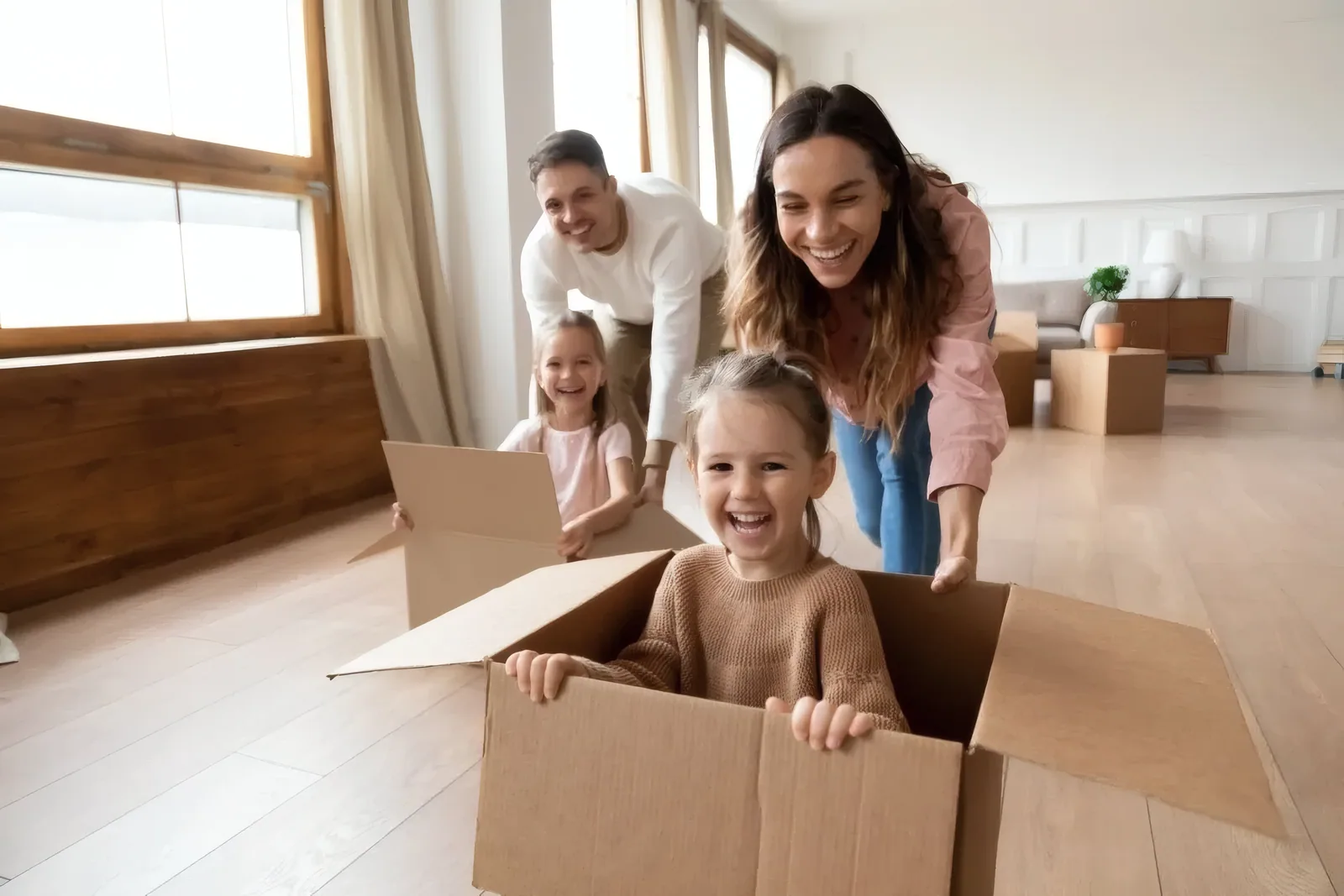 A joyful family playing in a bright, sunlit living room, parents pushing their laughing daughters along the floor in cardboard boxes.